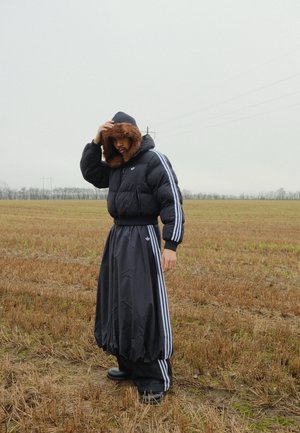 Chaqueta negra acolchada con capucha forrada de piel, combinada con una falda negra fluida con rayas blancas, llevada sobre pantalones oscuros y botas.
