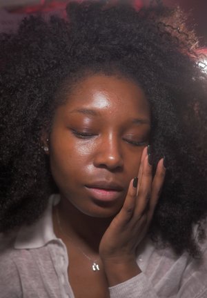 Joven con cabello rizado tocándose suavemente el rostro, con los ojos cerrados, vistiendo una camisa clara y un collar con un colgante.