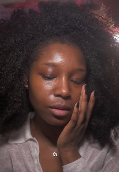 Joven con cabello rizado tocándose suavemente el rostro, con los ojos cerrados, vistiendo una camisa clara y un collar con un colgante.