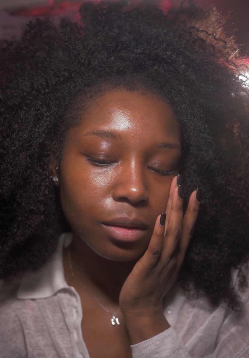 Jeune femme aux cheveux bouclés touchant doucement son visage, les yeux fermés, portant une chemise légère et un collier avec un pendentif.