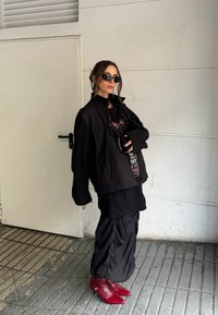 Černá oversized bunda s dlouhými rukávy, grafické tričko, vrstvená černá sukně a špičaté červené kotníkové boty, umístěné na světlejším pozadí.