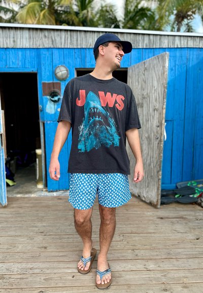 Hombre con camiseta negra de JAWS, pantalones cortos azules con estampado, gorra azul marino y sandalias caminando sobre un deck de madera cerca de un edificio azul con puertas abiertas.
