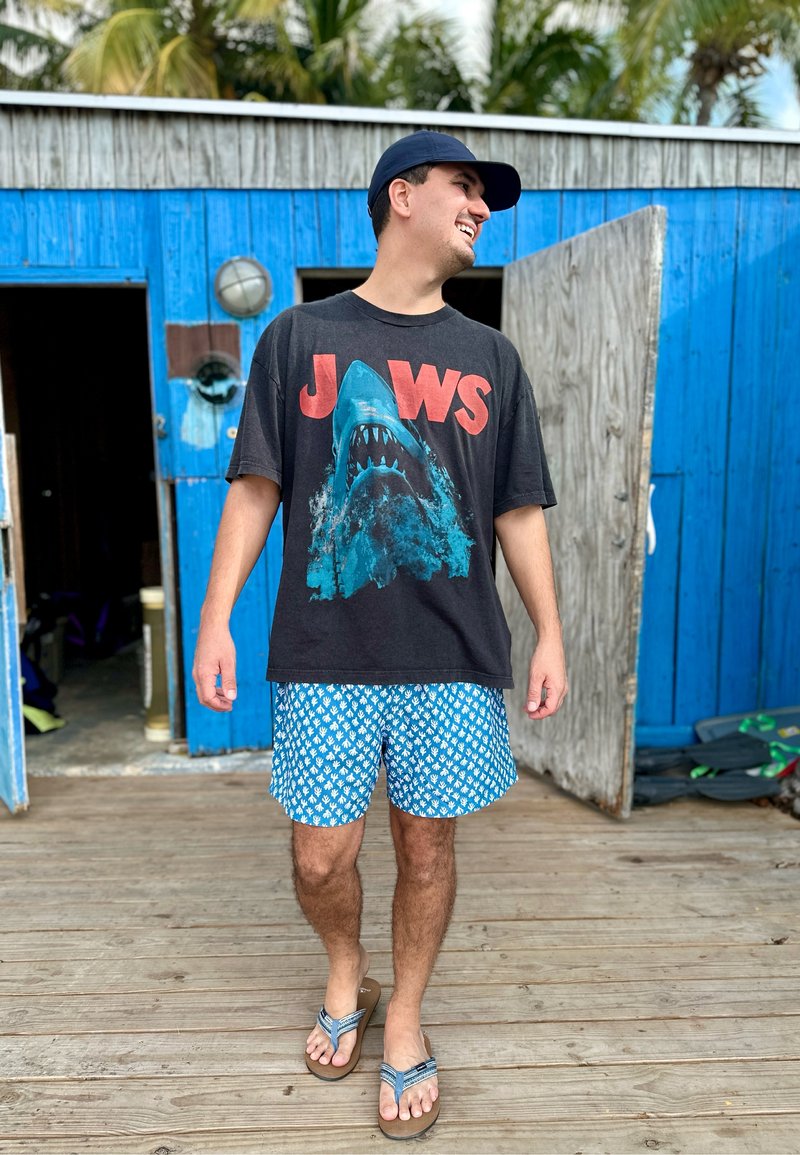 Vīrietis melnā T-kreklā ar "JAWS" uzrakstu, zilās rakstainās šortos, tumši zilā cepurē un sandales staigā pa koka terasi blakus zilai ēkai ar atvērtām durvīm.