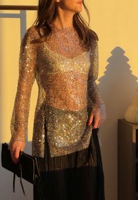 Femme portant un haut transparent à manches longues et paillettes sur un caraco, tenant une pochette noire, debout près de l'ombre d'un objet décoratif.