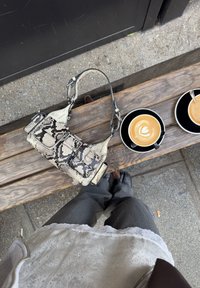 A snakeskin-patterned handbag in white and black rests on a wooden bench beside two coffee cups on black saucers. Grey trousers and a lace top are visible.