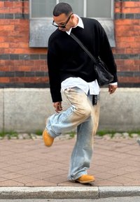 Sort strikket sweater, hvid skjorte under, lyseblå slidte jeans, gule træklip klip, sort crossbody taske, stående på en fortov.