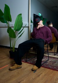 Braune Wanderschuhe, schwarze weite Hose und ein kastanienbrauner gerippter Pullover. Sitzend auf einem Stuhl neben einer großen grünen Pflanze und einem Spiegel.