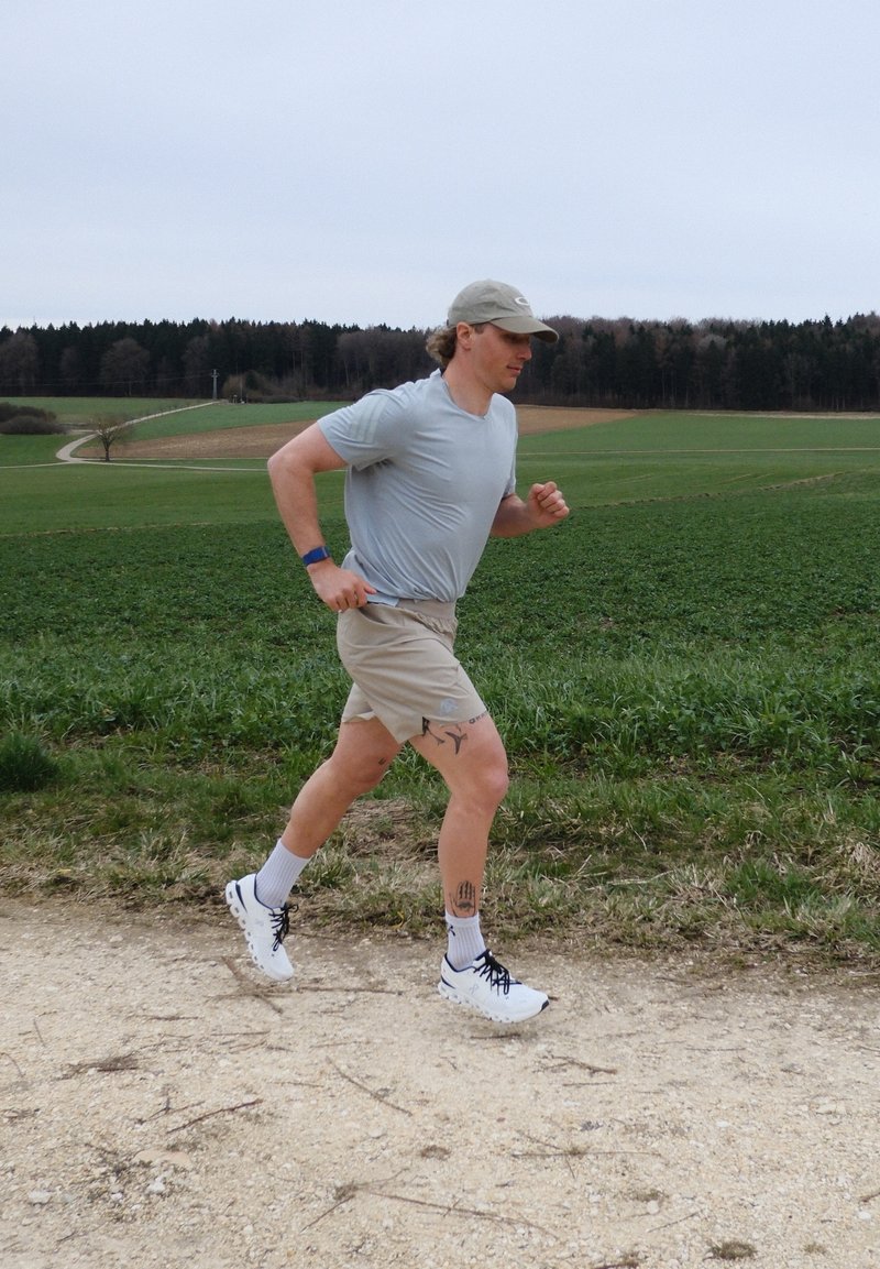 Man running on a gravel path through green fields under a cloudy sky, wearing light gray shirt, shorts, cap, white sneakers, and a fitness tracker.