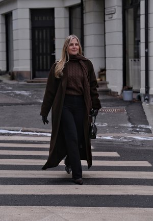 Mujer con abrigo oscuro, bufanda y guantes cruzando un paso de peatones en una calle de la ciudad, llevando un pequeño bolso negro.