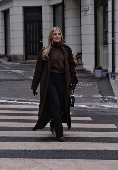 Mujer con abrigo oscuro, bufanda y guantes cruzando un paso de peatones en una calle de la ciudad, llevando un pequeño bolso negro.