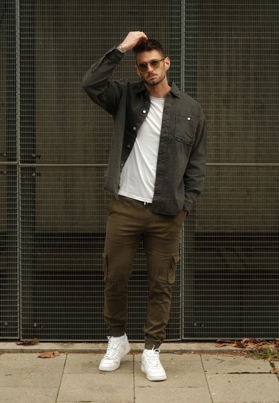 Camisa abotonada gris sobre una camiseta blanca, pantalones cargo oliva y zapatillas blancas. La persona lleva gafas de sol y el cabello despeinado.