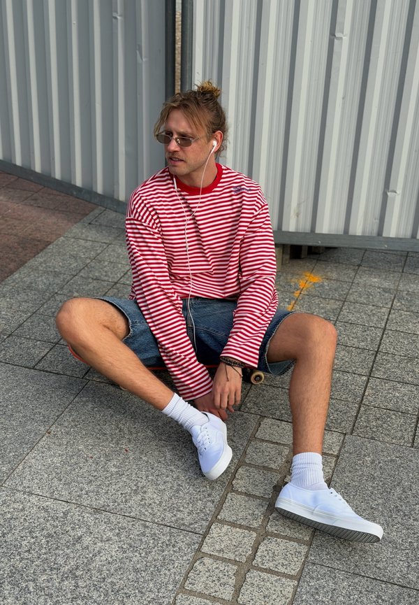 Joven con gafas de sol y auriculares, vestido con camiseta de rayas rojas y blancas y pantalones cortos de mezclilla, sentado con las piernas cruzadas en la acera con un monopatín.