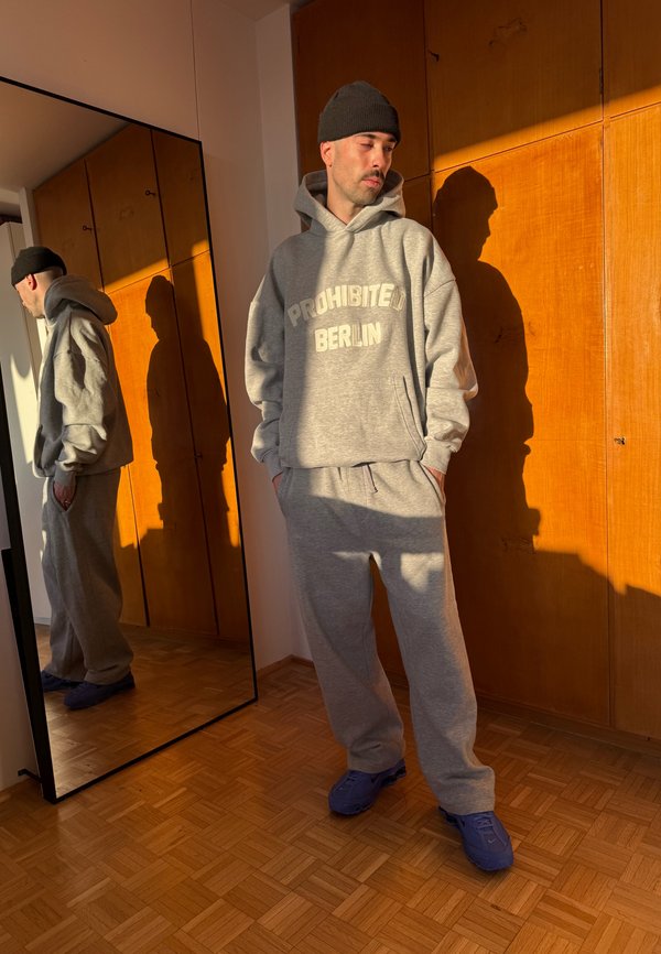 Man in een grijze "Prohibited Berlin" hoodie en sweatpants, zwarte beanie, blauwe schoenen, staand bij een spiegel en houten muur met warm zonlicht.