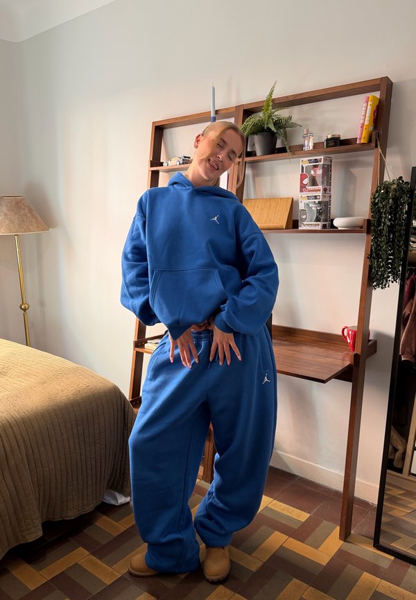 Jeune personne portant un survêtement bleu oversize et des bottes beige posant de manière ludique dans une chambre bien rangée avec des étagères en bois et un lit avec une couette marron.