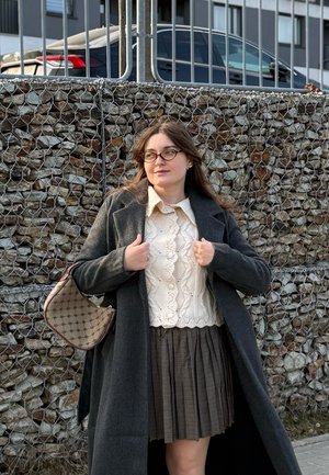 Joven con gafas que lleva un abrigo gris, un suéter de punto color crema y una falda plisada, de pie junto a una cerca de piedra con alambre, con coches aparcados detrás.