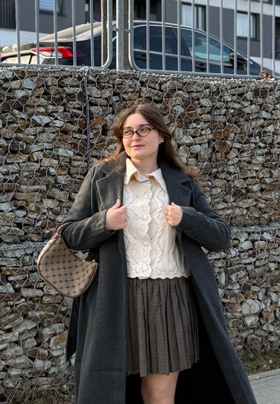 Joven con gafas que lleva un abrigo gris, un suéter de punto color crema y una falda plisada, de pie junto a una cerca de piedra con alambre, con coches aparcados detrás.