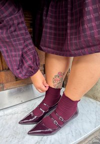 Sjajni burgundski Mary Jane cipele s dva remena i kopčama, uparene s maroon čarapama do gležnjeva. Pozadina sadrži drvene i kamenite teksture.
