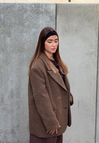 Blazer doppiopetto marrone testurizzato e oversize con grandi rever e bottoni marroni, indossato sopra una maglia scura, su sfondo di cemento.