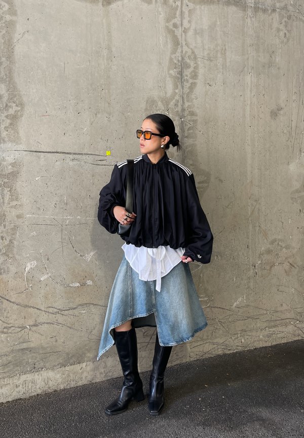 Mujer con chaqueta negra de gran tamaño, camisa blanca en capas, falda vaquera asimétrica, botas negras, gafas de sol naranjas, de pie contra una pared de hormigón.