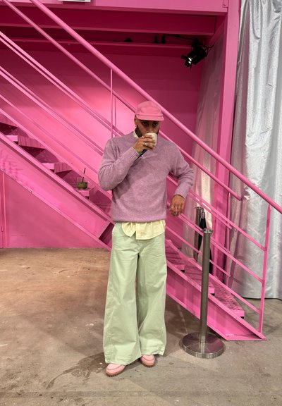 Gorra rosa claro, suéter lavanda, camisa color crema y pantalones anchos verde menta, de pie sobre un suelo gris con una escalera rosa en el fondo.