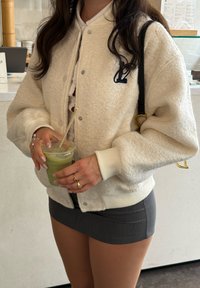 Weißer Fleece-Bomberjacke mit gerippten Bündchen und Saum, mit einem marineblauen Buchstabenlogo. Grauer Rock. Hält einen transparenten Becher mit grünem Getränk.