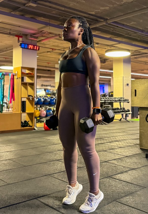 Nő sportfelszerelésben, súlyzókat emel egy edzőteremben, gumírozott padlón állva, mögötte kettlebellek és súlyzók láthatók.