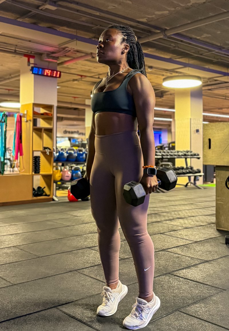 Kobieta w stroju sportowym podnosząca hantle na siłowni, stojąca na gumowej podłodze, w tle kettlebelle i ciężary.