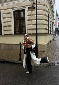 Femme en manteau blanc et lunettes de soleil tenant un bouquet festif, debout à un coin de rue mouillé près d'un bâtiment orné et d'un lampadaire noir.