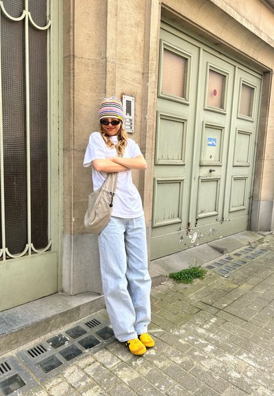 Conjunto casual que incluye una camiseta blanca, jeans de pierna ancha en azul claro, zapatillas amarillas y un gorro a rayas multicolor. Un bolso cruzado beige completa el look.