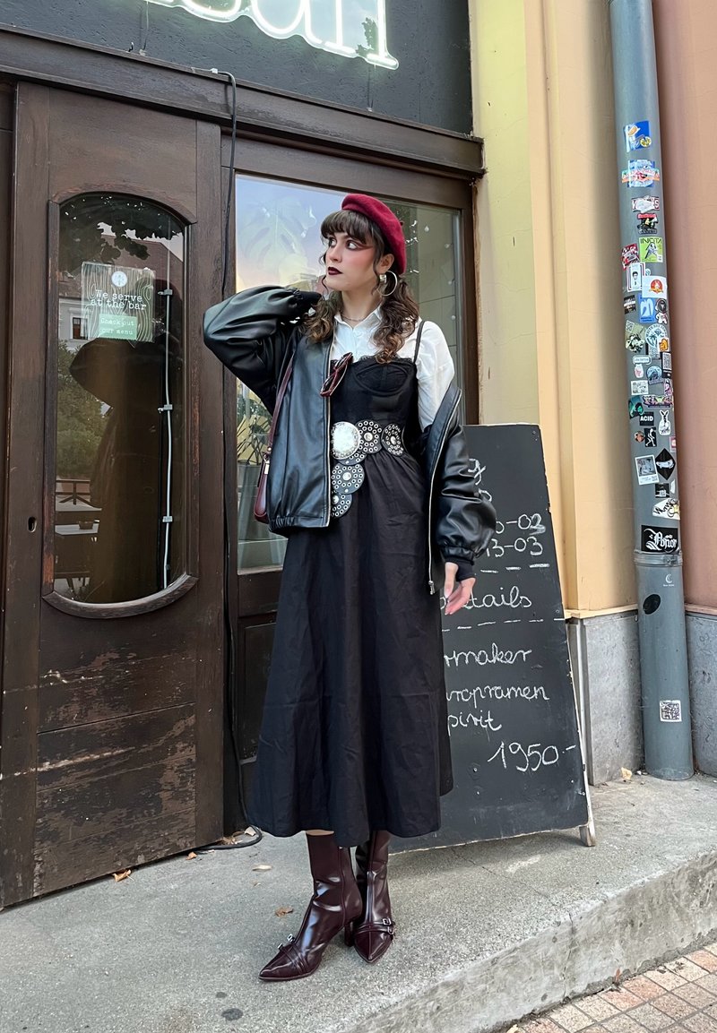 Vestido negro midi adornado con embellishments circulares plateados, superpuesto a una camisa blanca, acompañado de una chaqueta bomber negra y botas de tacón burdeos.