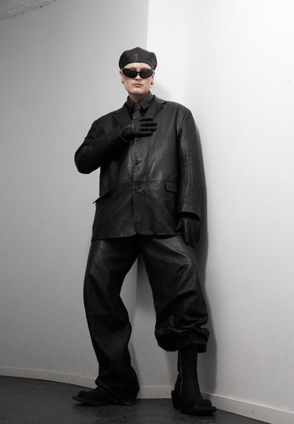 Person iført oversized sort læderjakke, bukser, handsker, baret og solbriller, stående mod et hvidt hjørnevæg.