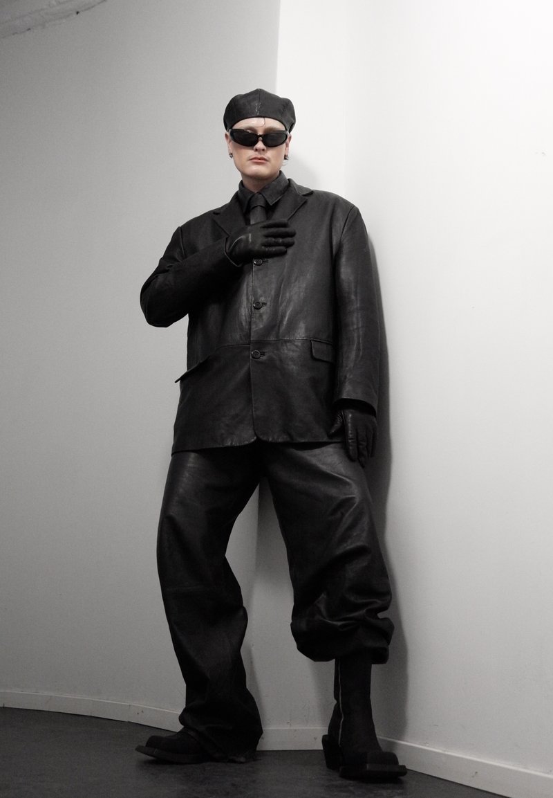 Personne portant une veste en cuir noire surdimensionnée, un pantalon, des gants, un béret et des lunettes de soleil, se tenant contre un mur blanc d'angle.
