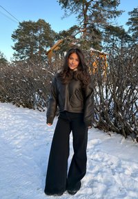 Femme debout sur un sol enneigé, portant une veste en cuir noire et un pantalon large noir, avec des arbres et une maison en arrière-plan.