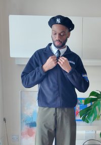 A man wearing a black Adidas jacket, a white shirt, a black tie, and a navy beret is adjusting the zipper of his jacket in a modern room adorned with plants and artwork.