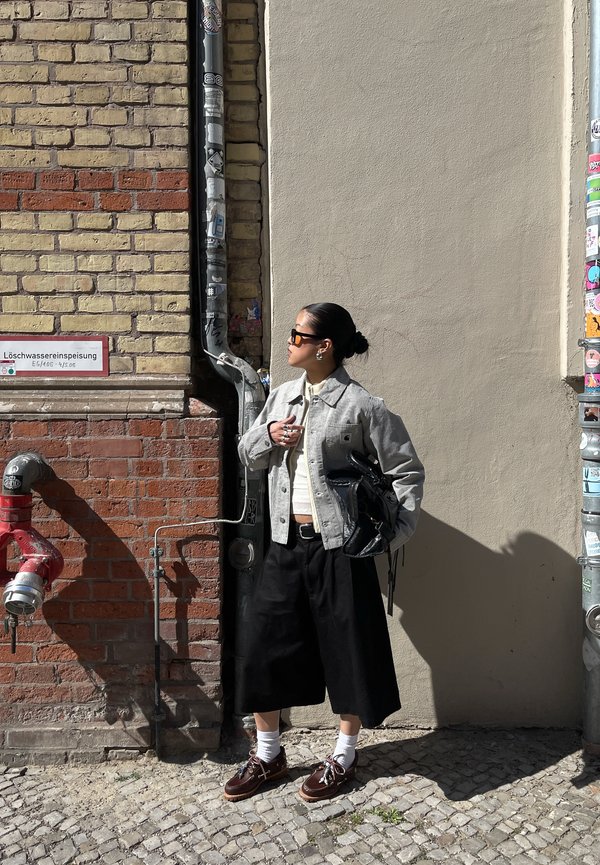 Nő napszemüvegben, konty hajjal, egy téglából és vakolatból álló városi falnak támaszkodva, vörös vízcsővel és matricákkal borított oszloppal.