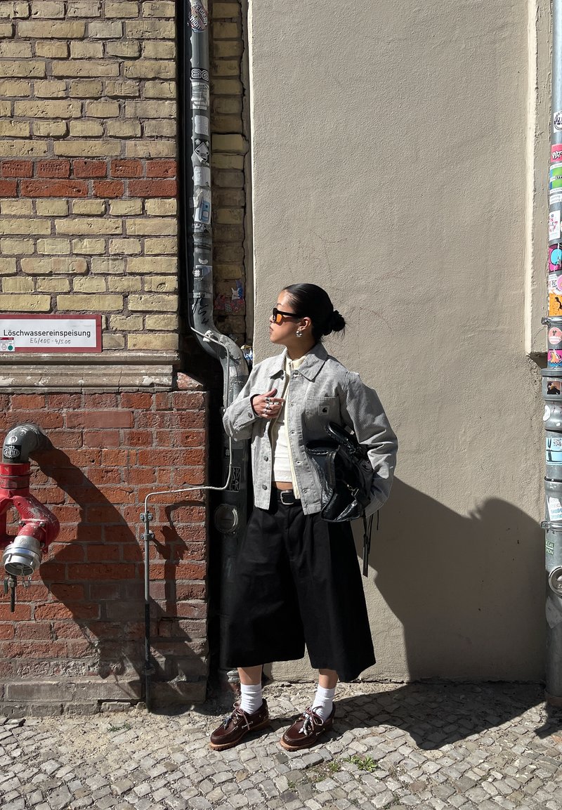 Femme avec des lunettes de soleil et un chignon, debout contre un mur urbain en briques et plâtre avec un tuyau d'eau rouge et un poteau couvert d'autocollants.