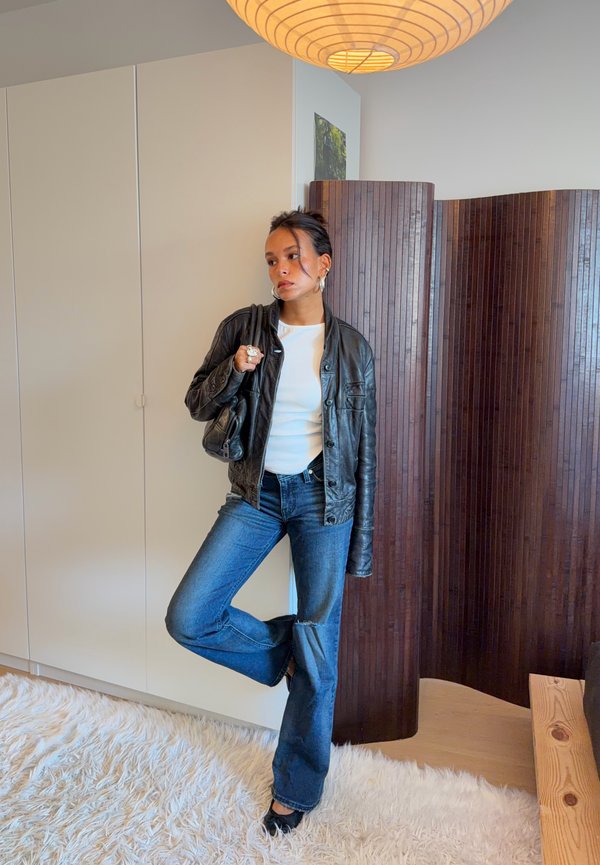 Una mujer con una chaqueta de cuero negra, una camiseta blanca y vaqueros posa junto a un biombo de madera en una habitación con una iluminación cálida.