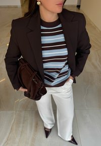 Blazer marrone sopra un maglione a righe blu e marrone, pantaloni bianchi a gamba larga e scarpe scure con punta. Borsa a tracolla marrone scuro con cinturino.