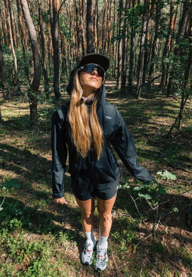 Femme portant une veste noire, un short, une casquette et des lunettes de soleil, debout dans une forêt ensoleillée avec de grands arbres et un sous-bois vert.