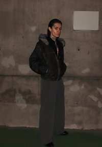 Črni usnjeni jopič z ovratnikom iz krzna, sive nabrane hlače, stoji ob betonskem zidu. Tetovaže vidne na vratu. Črni čevlji.