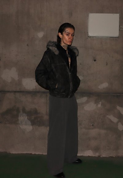 Chaqueta de cuero negro con cuello de piel, pantalones plisados grises, de pie contra una pared de concreto. Tatuajes visibles en el cuello. Zapatos negros.