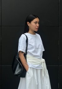 Witte katoenen t-shirt en rok met een gestrikte witte sjerp. Zwarte leren schoudertas. Model met lang, donker haar en creolen.