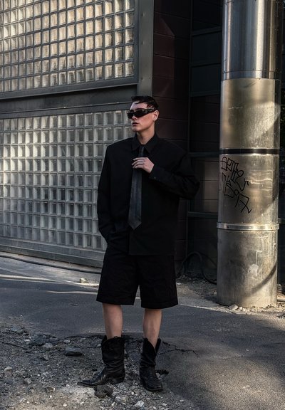 Camisa negra de gran tamaño, pantalones cortos a juego, corbata negra y botas altas negras. Los accesorios incluyen gafas de sol con montura única. Fondo industrial.