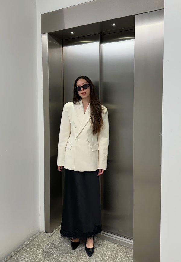 Kvinne iført hvit blazer, lang svart skjørt, svarte hæler og solbriller står foran lukkede sølvfargede heisdører innendørs.