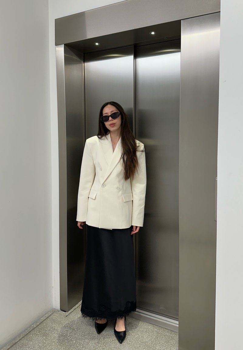 Donna con blazer bianco, gonna lunga nera, tacchi neri e occhiali da sole in piedi davanti a porte d'ascensore argento chiuse all'interno.