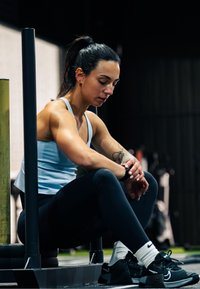 Tenue de sport comprenant un haut sans manches bleu clair et un legging noir. Femme assise, avec un tatouage sur le bras et portant des baskets noires.