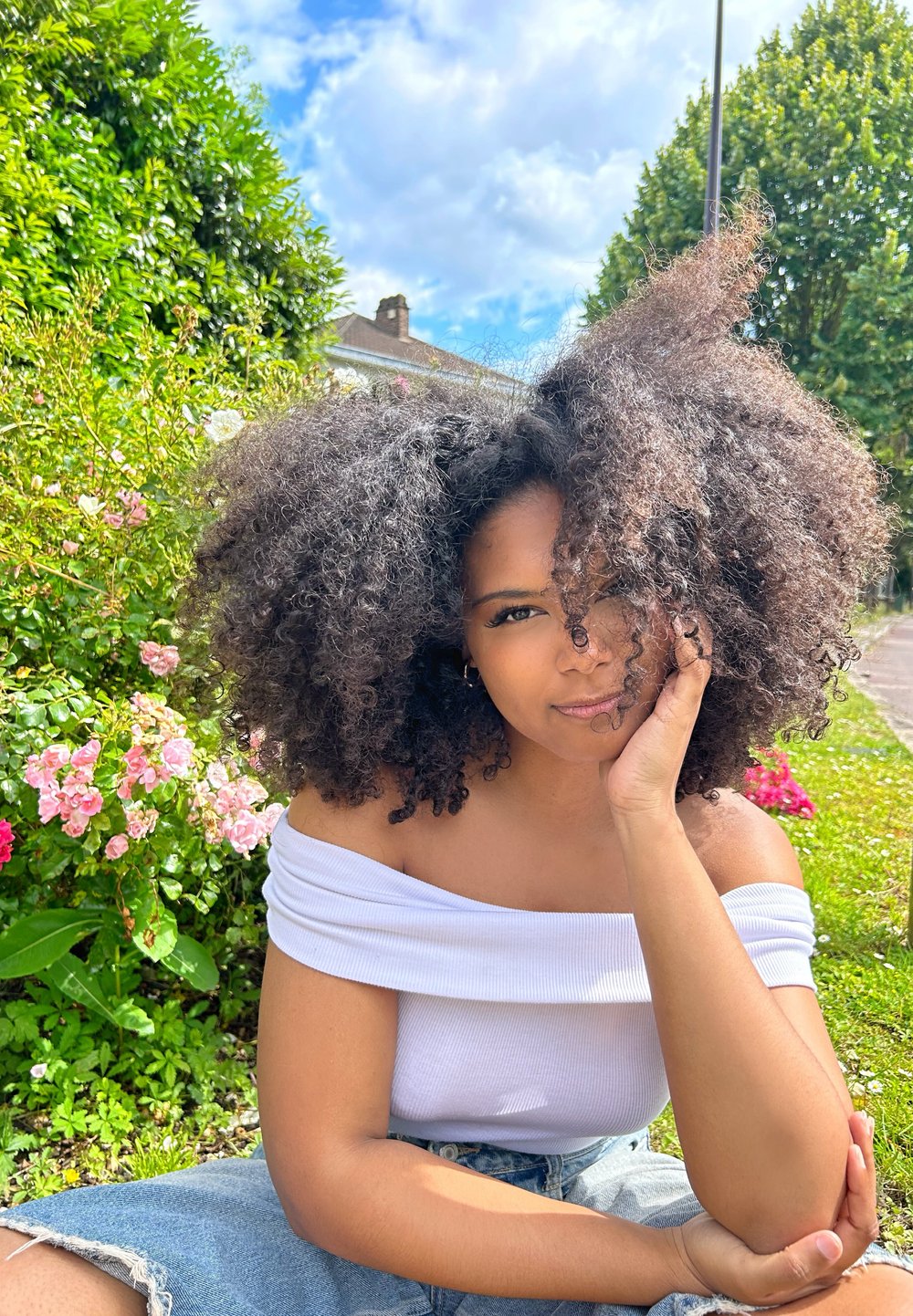 Ein Model mit voluminösen schwarzen Locken sitzt auf dem Gras, trägt eine weiße schulterfreie Bluse und helle Denim-Shorts, mit pinken Blumen im Hintergrund.