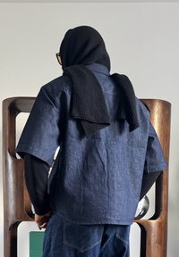 Personne portant des vêtements en denim superposés et noirs avec une écharpe tricotée noire drapée sur la tête et les épaules, debout devant des étagères en bois.