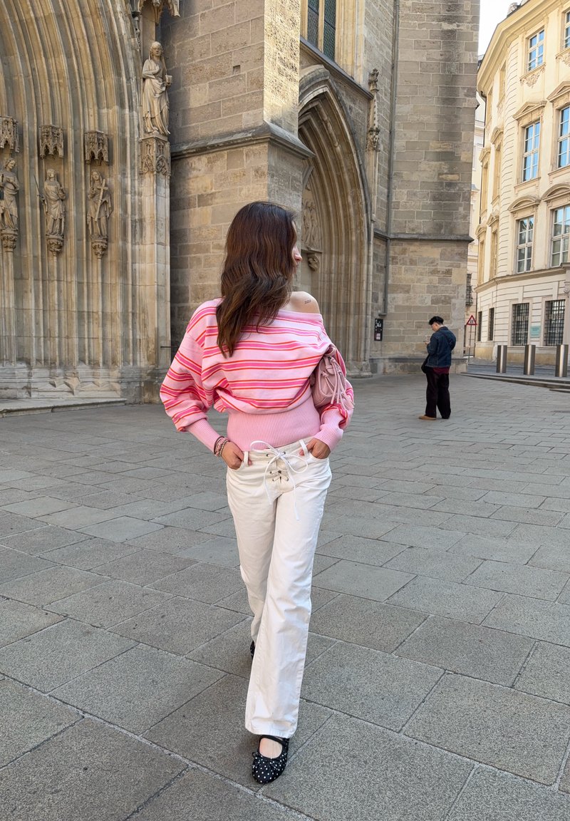 Woman in pink striped off-shoulder sweater and white pants walking on stone pavement near a Gothic stone building with statues.