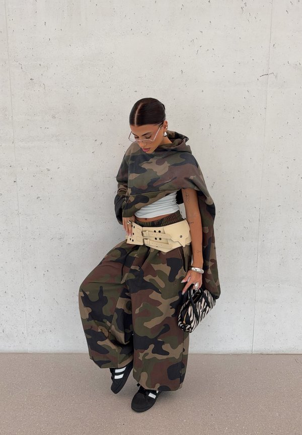 Mujer con pantalones anchos de camuflaje y sudadera con capucha con cinturón beige, zapatillas negras, gafas transparentes y sosteniendo un bolso con estampado de cebra contra una pared lisa.